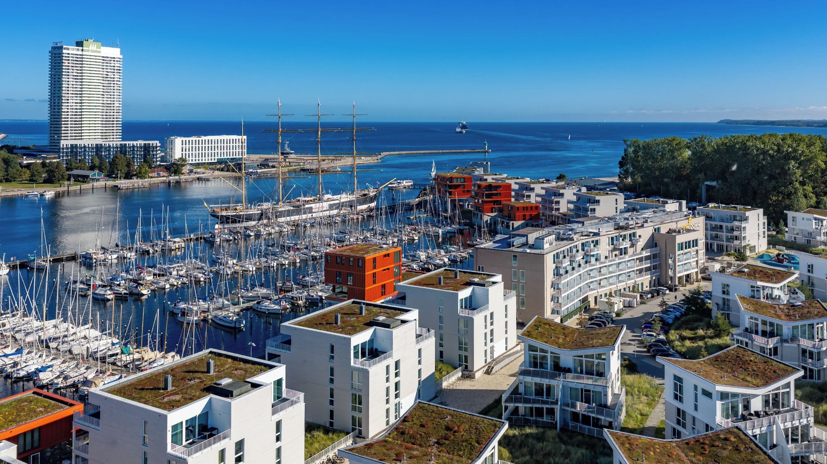 Un port de plaisance avec de nombreux bateaux, des bâtiments modernes dotés de toits plats et une vue dégagée sur la mer.