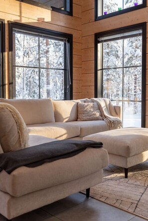 A modern living room with a sofa and ottoman, large windows with a snowy view, and wooden paneling on the walls. - Stay North - Levin Sevilla (Kittilä)