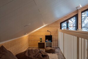 A cozy attic room with a sloped ceiling, wooden walls, a small seating area with cushions, a television, and a window with a view of trees. - Stay North - Levin Sevilla (Kittilä)