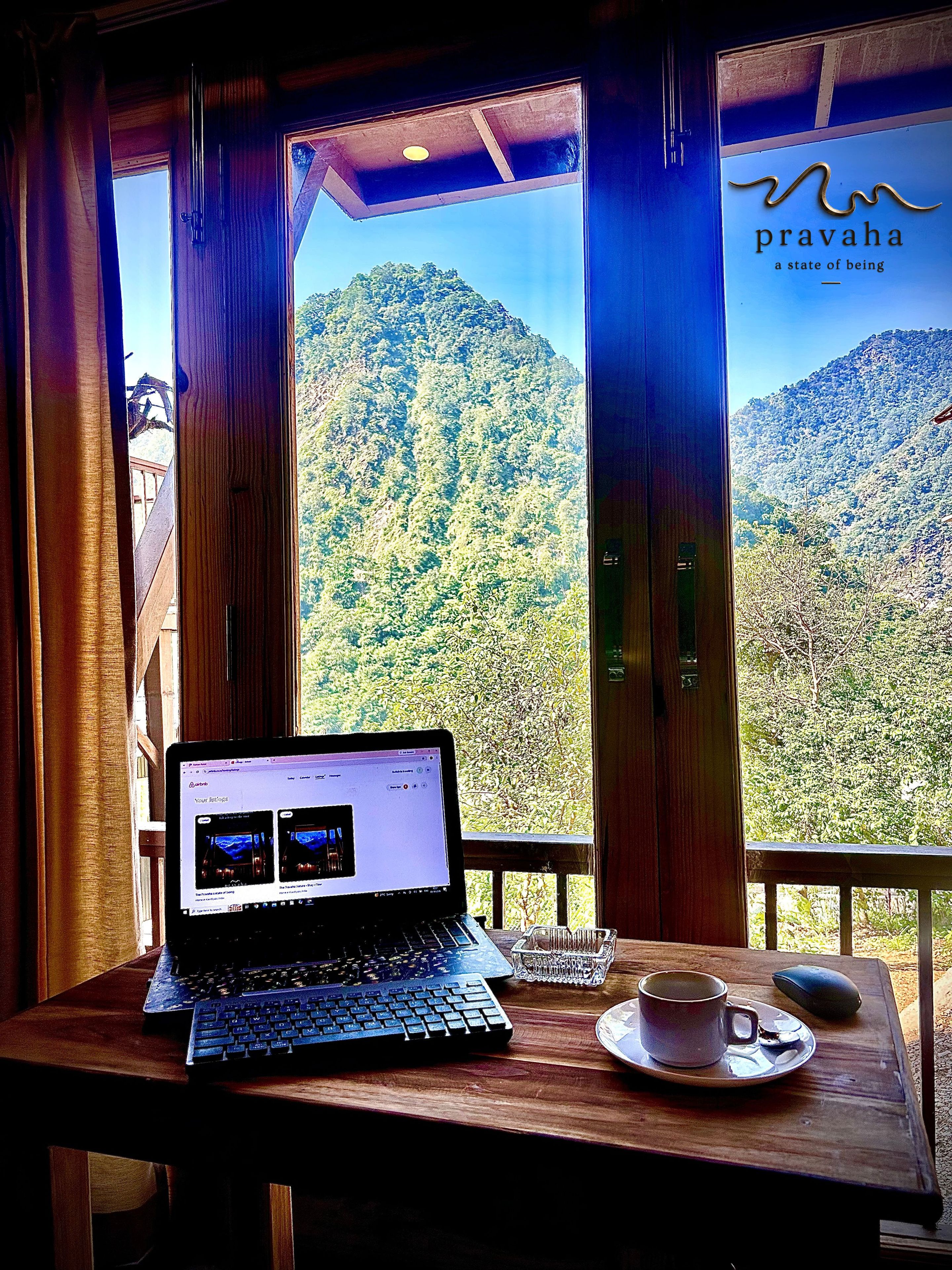 Una computadora portátil sobre una mesa de madera con una taza y platillo, un ratón y un teclado. Una ventana muestra un paisaje montañoso.