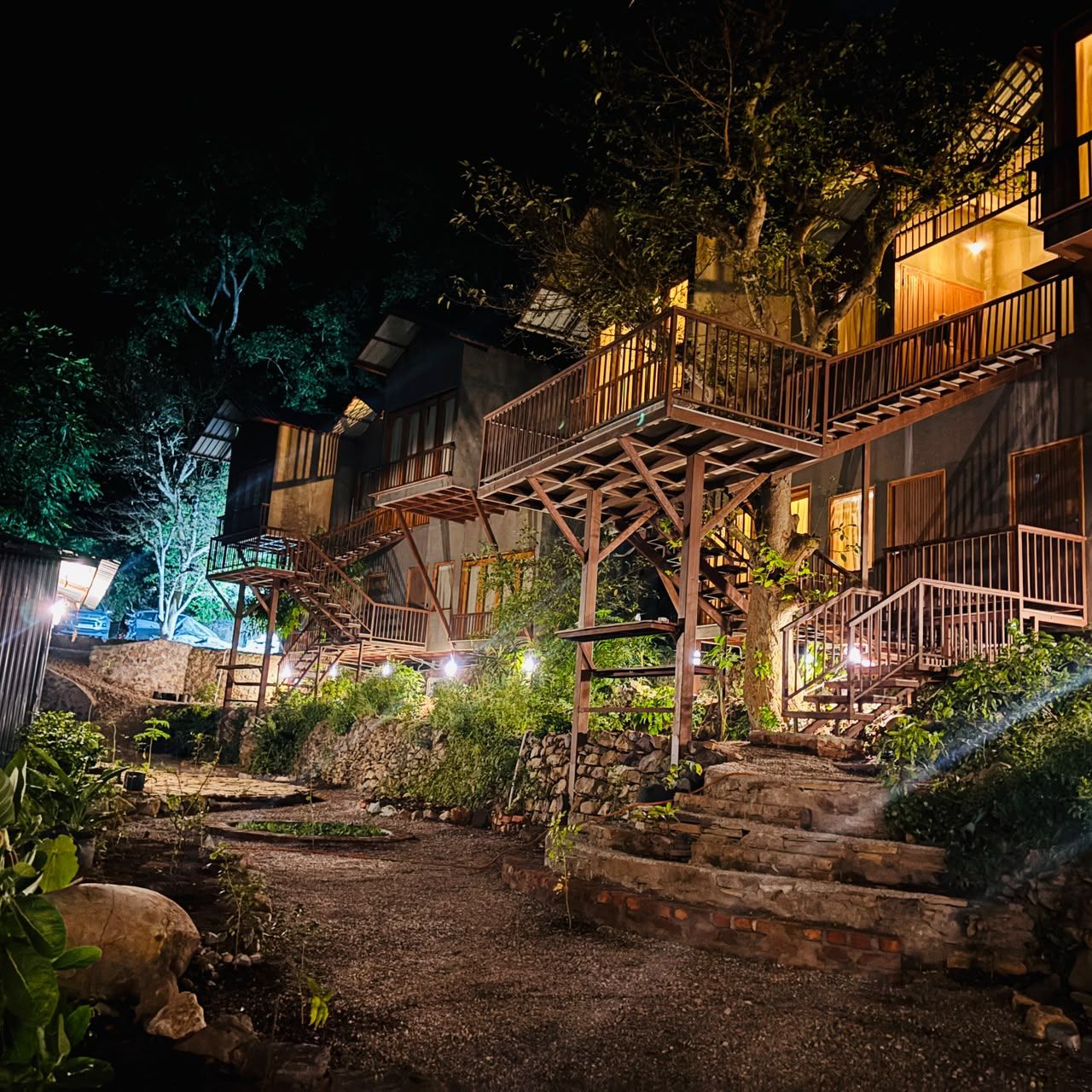 A multi-level wooden structure with outdoor stairs, surrounded by greenery and stone walls.