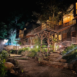 A multi-level wooden structure with outdoor stairs, surrounded by greenery and stone walls.