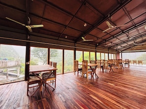 A spacious indoor area with wooden flooring, multiple tables and chairs, and large windows offering a view of the outdoors.