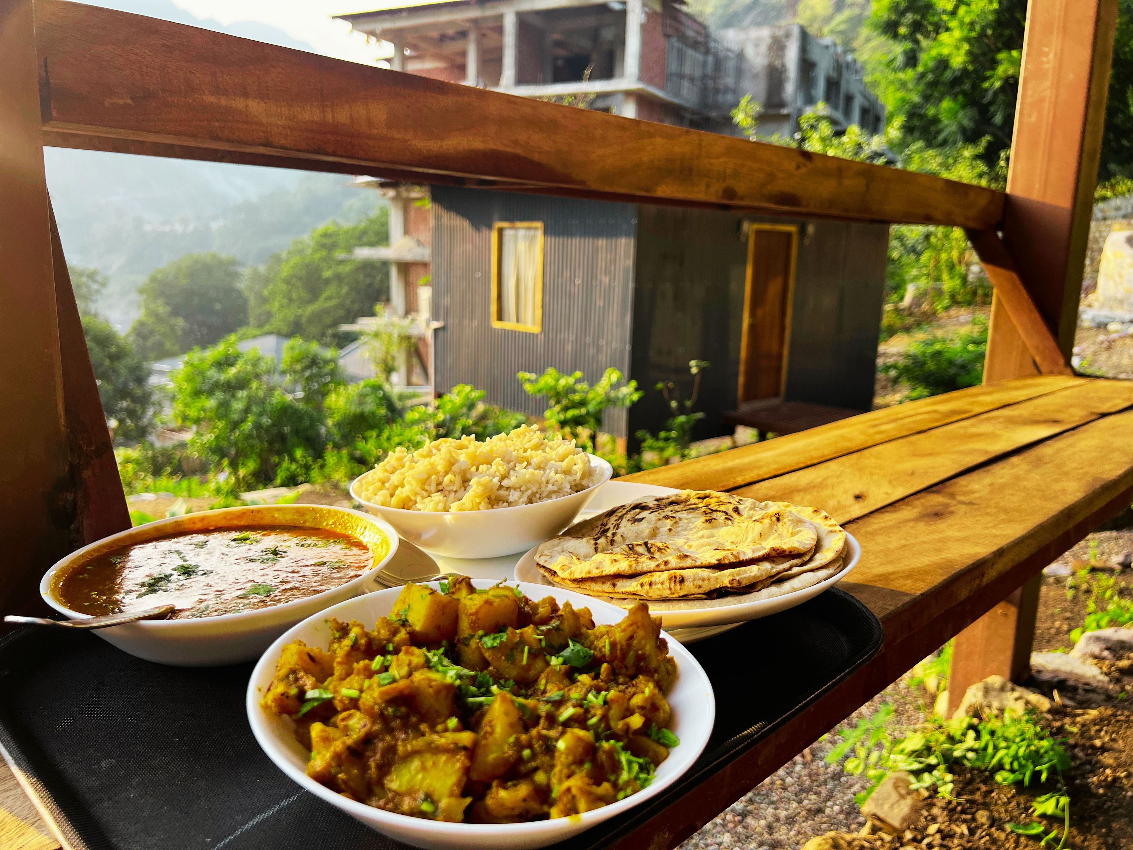 A wooden bench with a tray of food including rice, curry, and flatbreads, overlooking a residential area with greenery and buildings.