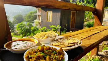A wooden bench with a tray of food including rice, curry, and flatbreads, overlooking a residential area with greenery and buildings.