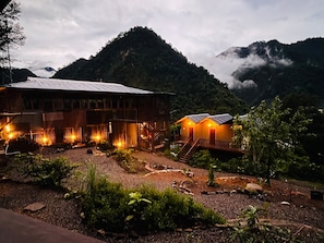 Un chalet rustique doté d’un grand bâtiment en bois et d’une structure plus petite avec un toit en pente, le tout entouré d’un jardin aménagé et surplombé par un paysage montagneux.