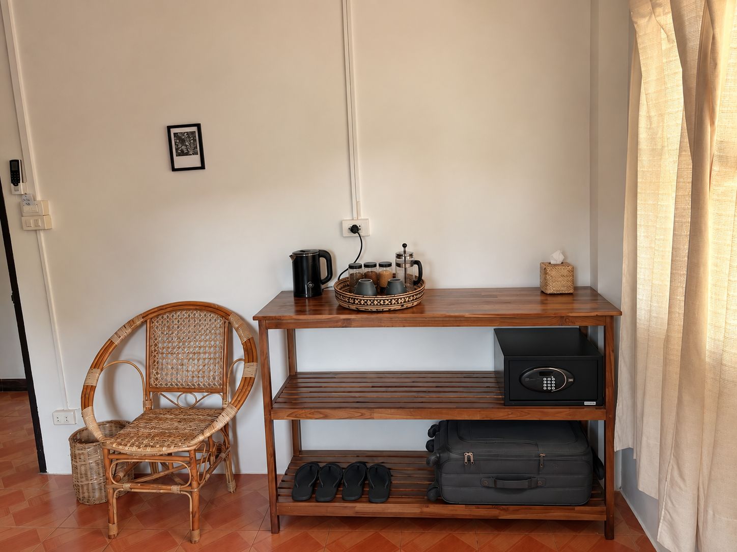 Ein Zimmer mit einem Korbstuhl, einem Holztisch mit einer Auflage und verschiedenen Gegenständen, einem schwarzen Koffer und einem schwarzen Lautsprecher.