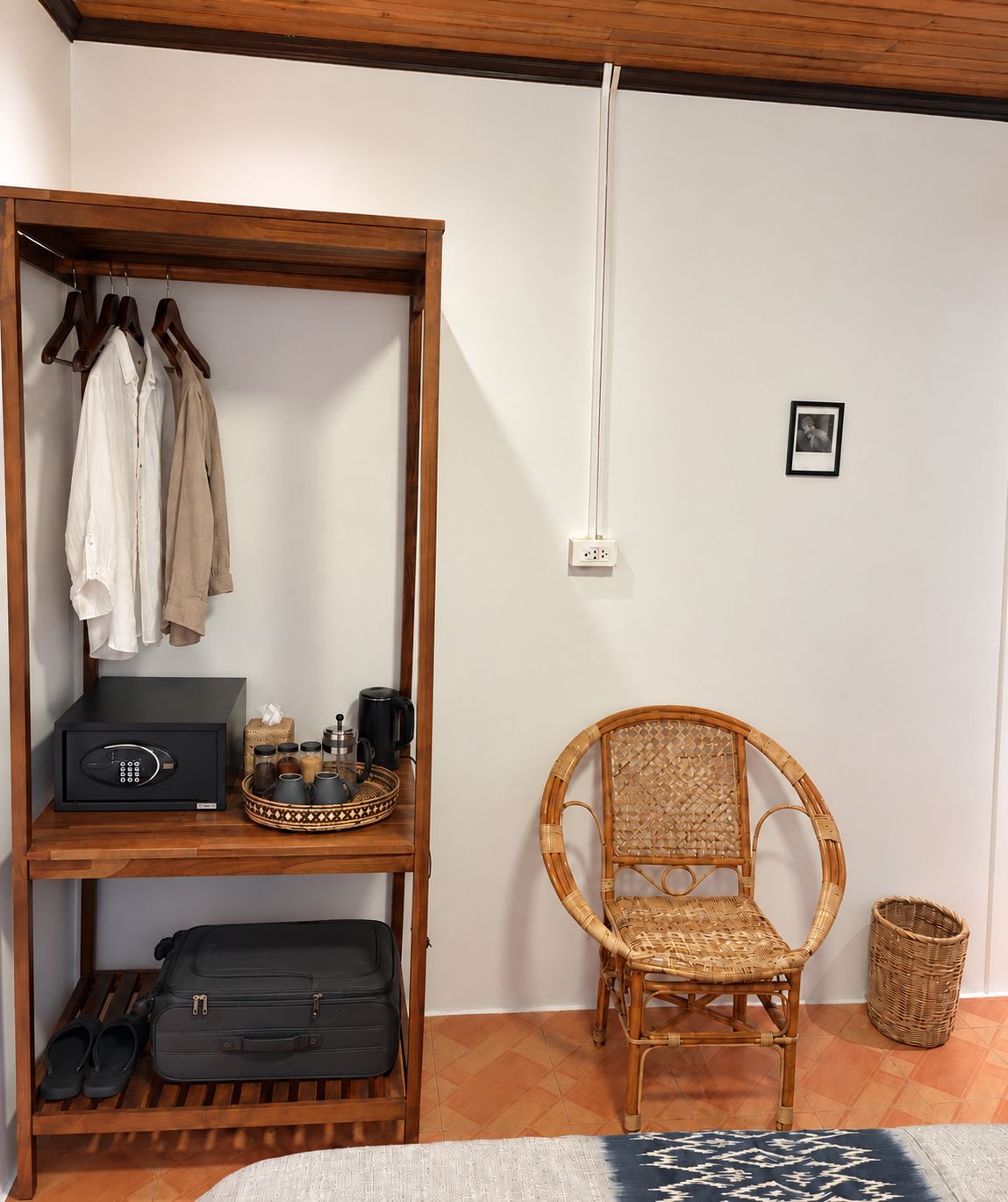 Une pièce avec une étagère en bois où sont rangés des vêtements, une radio noire et un plateau contenant des ustensiles de cuisine. On y trouve également une chaise en osier et un panier tressé posés sur le sol.