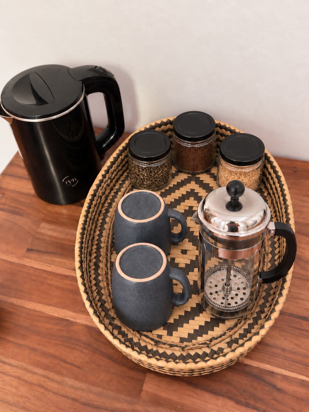Ein Kaffee-Arrangement mit einer schwarzen Kaffeemaschine, zwei dunklen Tassen, einer French Press und drei Gewürzbehältern auf einem gewebten Tablett.