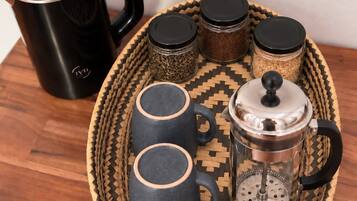 Un ensemble pour préparer le café comprenant une cafetière noire, deux tasses foncées, une cafetière à piston et trois pots à épices, le tout disposé sur un plateau en osier.