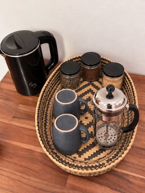 A coffee setup with a black coffee maker, two dark mugs, a French press, and three spice jars on a woven tray. - Heuan Xieng Thong (Luang Prabang)