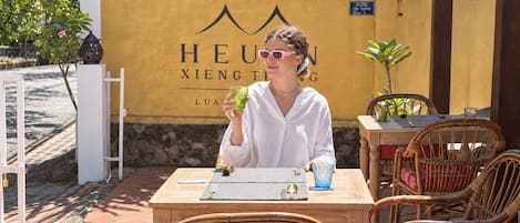 A person seated at an outdoor table with a drink, surrounded by wicker chairs and a yellow wall with signage.