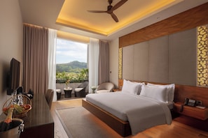 Habitación de hotel con una cama grande, un escritorio con una botella de champán, una silla, una mesita con un florero y vistas a zonas verdes.