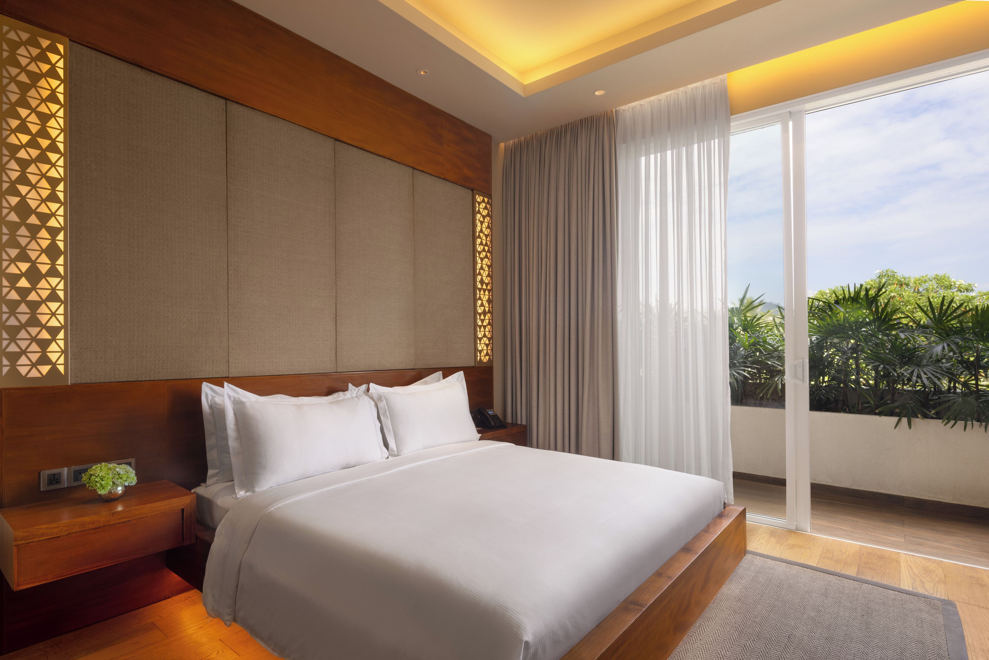 A modern hotel room with a large bed, a wooden headboard, a bedside table with a phone, and a view of greenery through the window.