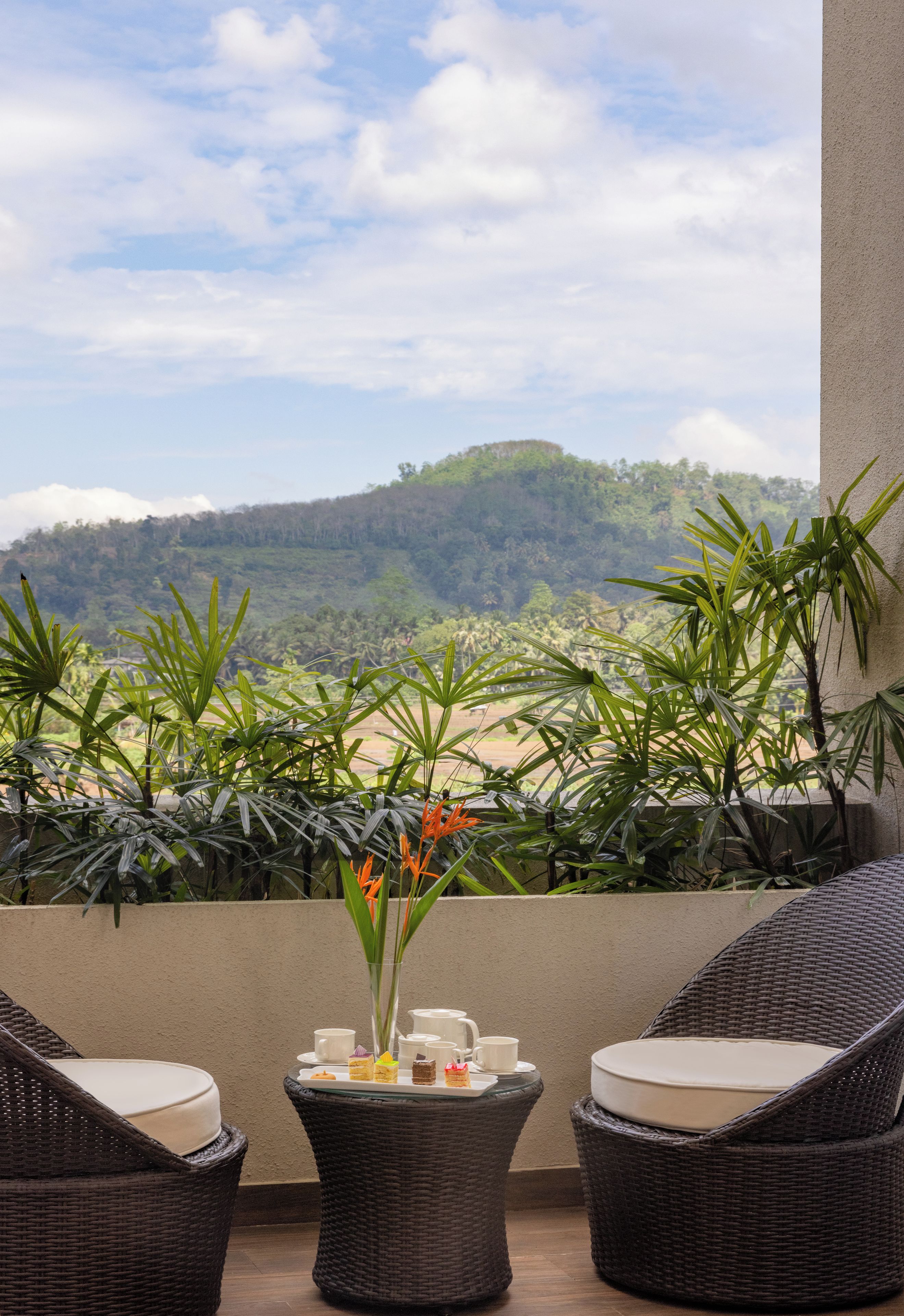 Un balcon agrémenté de meubles en osier, une petite table avec un vase de fleurs et une vue sur une colline boisée.