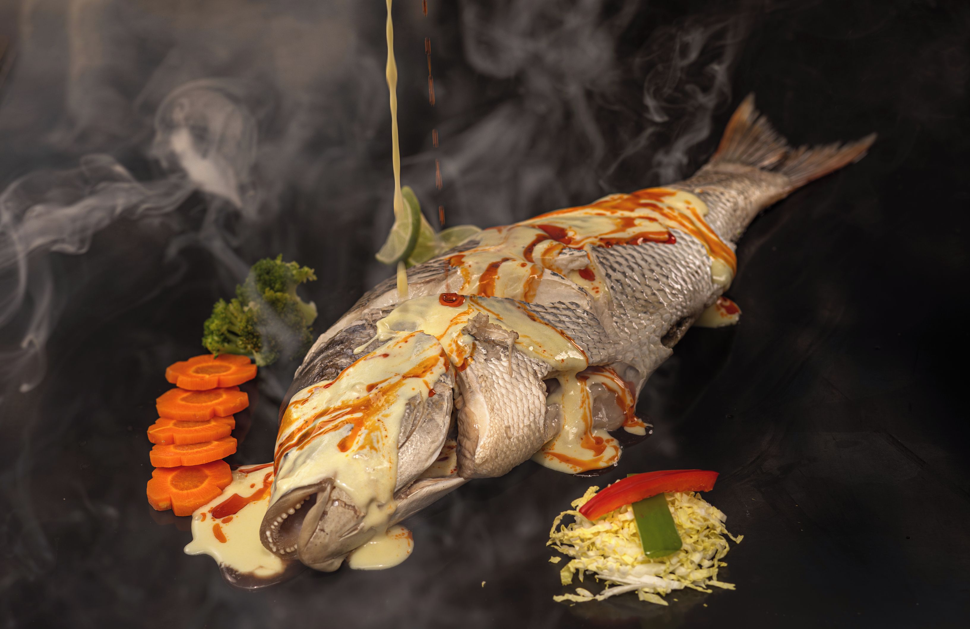 A whole fish with sauce and garnishes on a black surface.