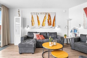 A modern living room with a grey sectional sofa, a wooden coffee table, and a painting of sailboats on the wall. - Spacious holiday apartment with luxury features and covered terrace near the Bal (Lübeck)