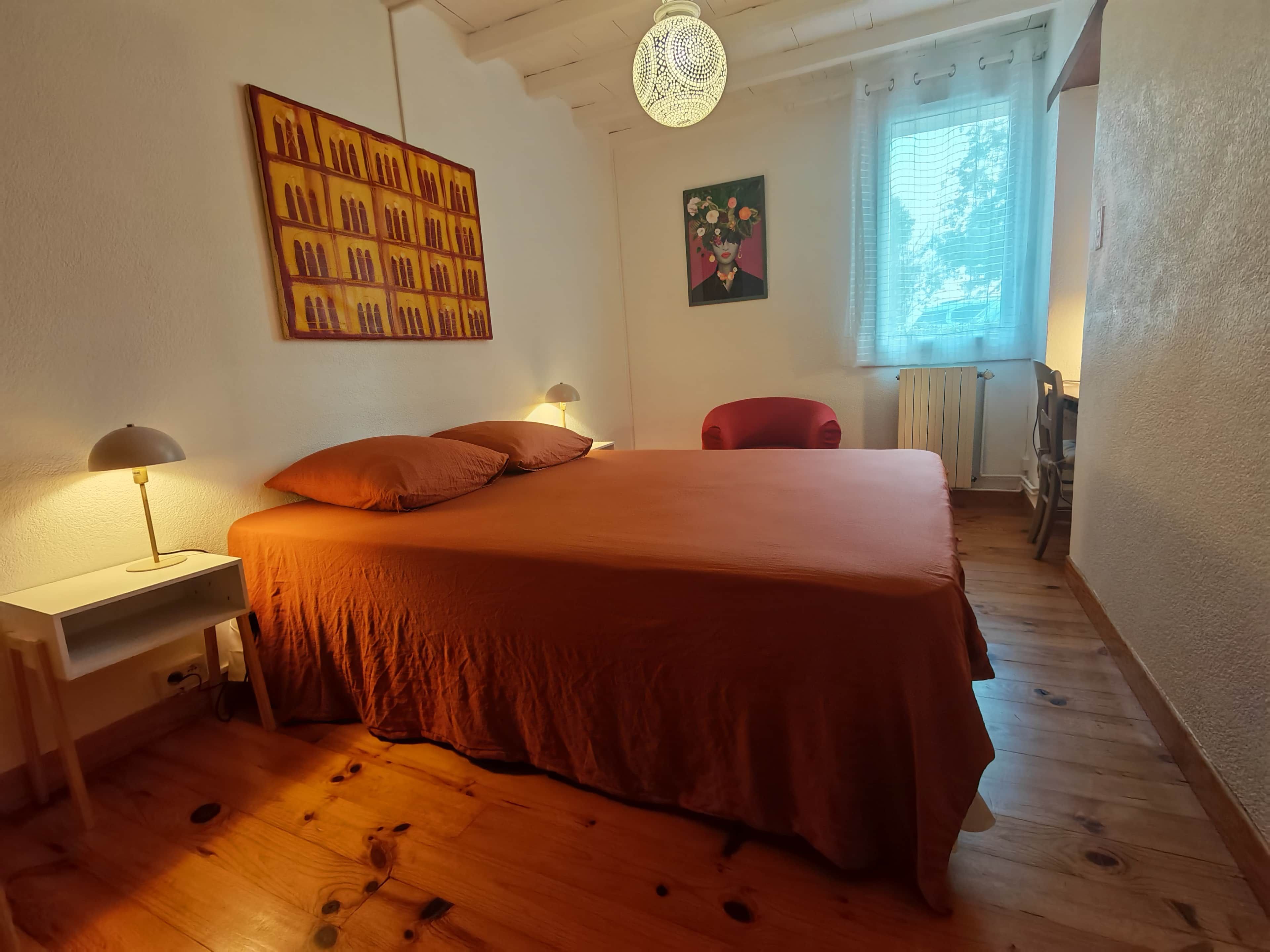 Une chambre à coucher avec un grand lit, une table de chevet avec une lampe, une chaise, un tableau au mur et une fenêtre avec des rideaux.