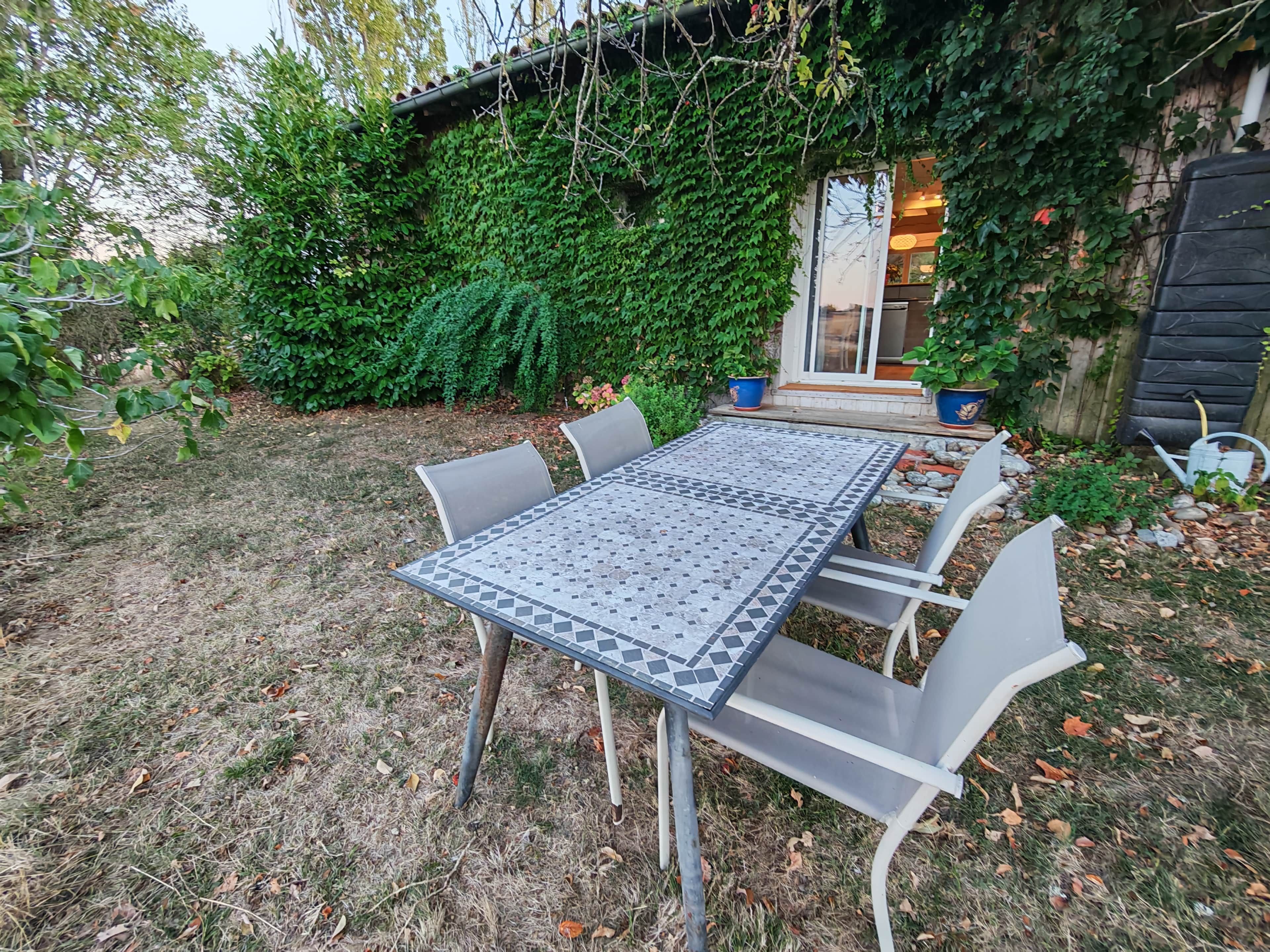 Une terrasse extérieure avec une table et des chaises carrelées, entourée de verdure et d’un bâtiment.