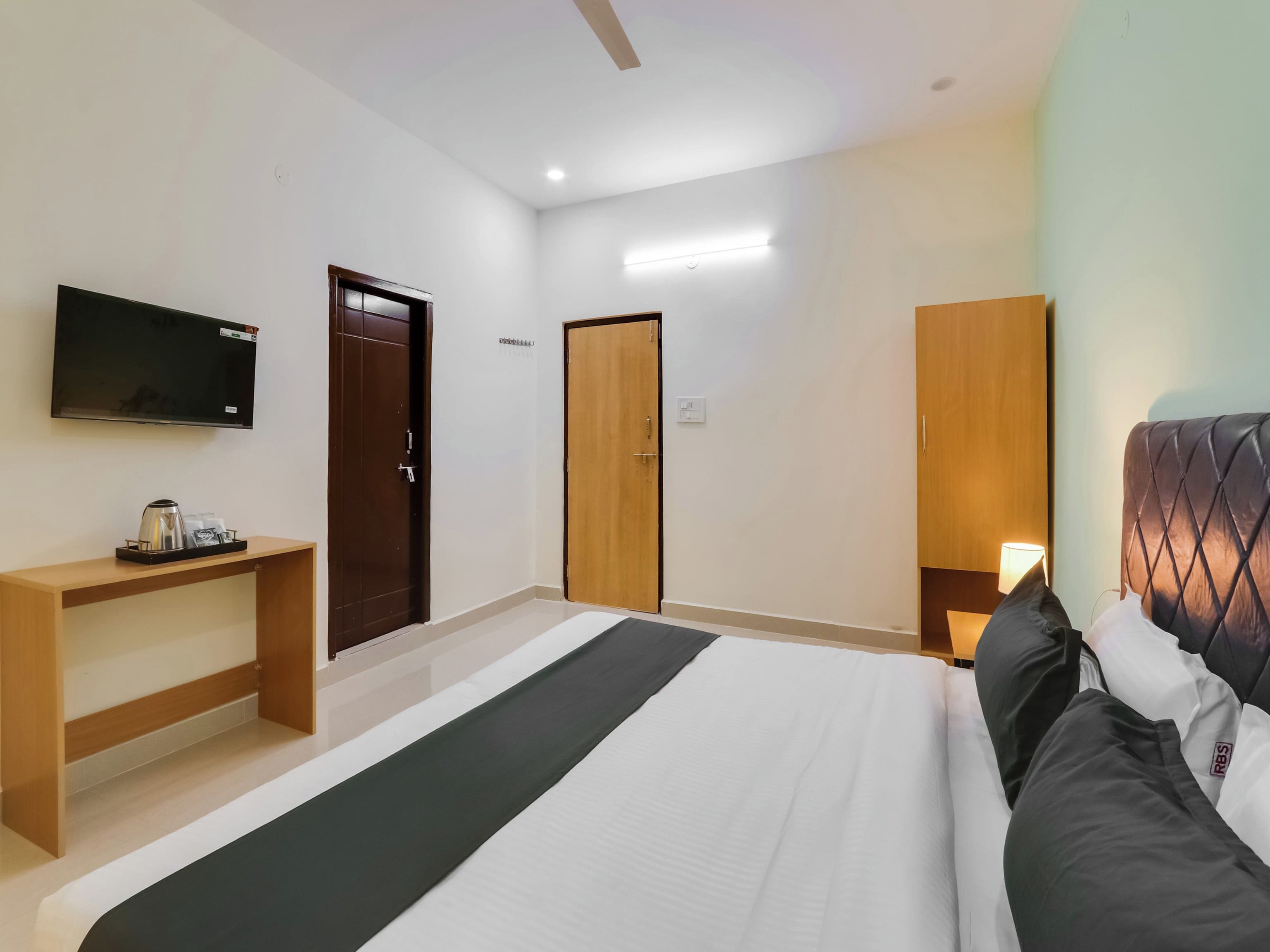 A hotel room with a bed, bedside table, TV, wardrobe, and a desk with a kettle.