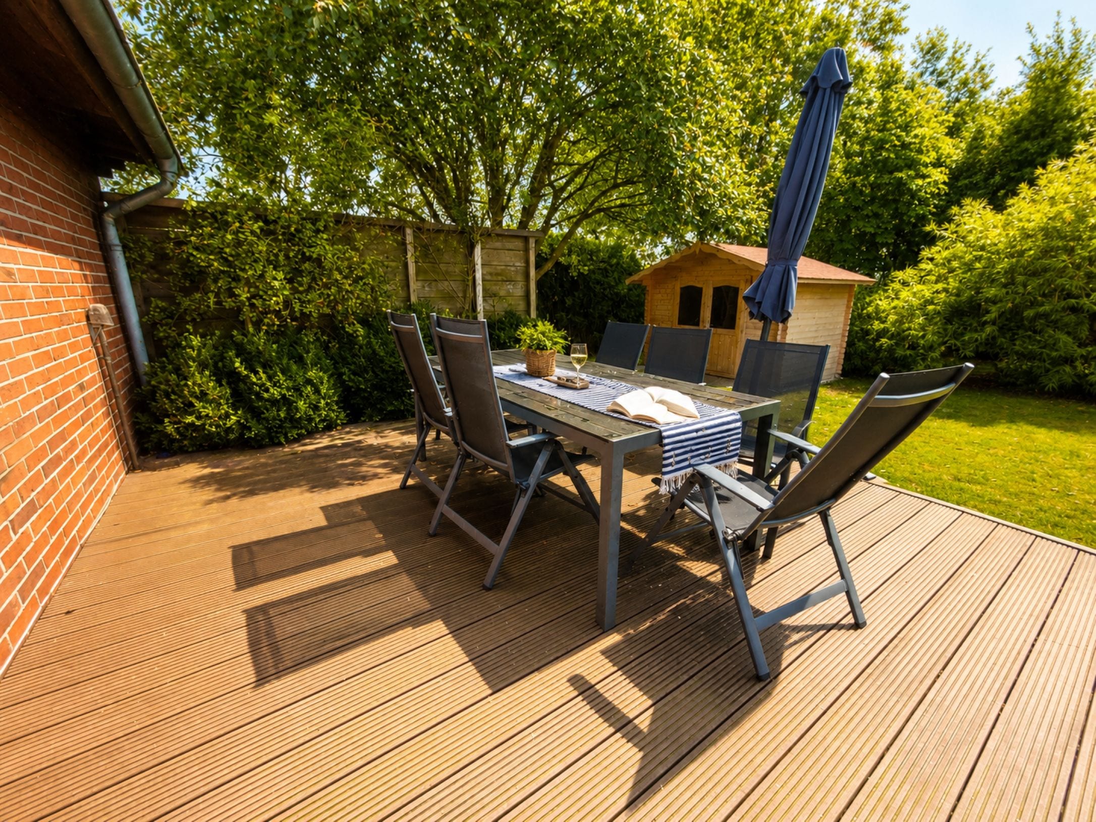 Una terraza de madera con una mesa y sillas, rodeada de follaje y una pequeña construcción de madera.