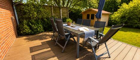 Una terraza de madera con una mesa y sillas, rodeada de follaje y una pequeña construcción de madera.