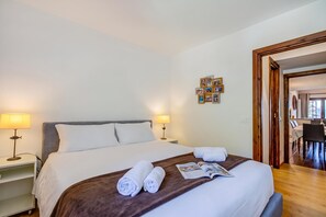 Habitación de hotel con cama, dos lámparas de noche, un collage de fotos en la pared y un área de comedor visible a través de una puerta abierta.
