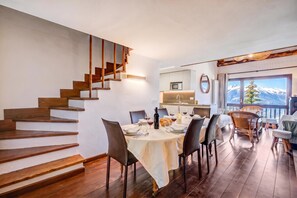 Un comedor con una mesa dispuesta para cuatro, una escalera con escalones de madera y vistas a montañas nevadas a través de una ventana.