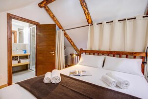 Habitación de hotel con cabecera de madera, cama con ropa blanca, alfombra tejida marrón y vista a un baño con espejo y lavamanos.