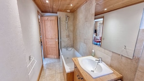A bathroom with a wooden door, a bathtub, a sink, and a mirror. - Apartment for rent near the city center. (Megève)