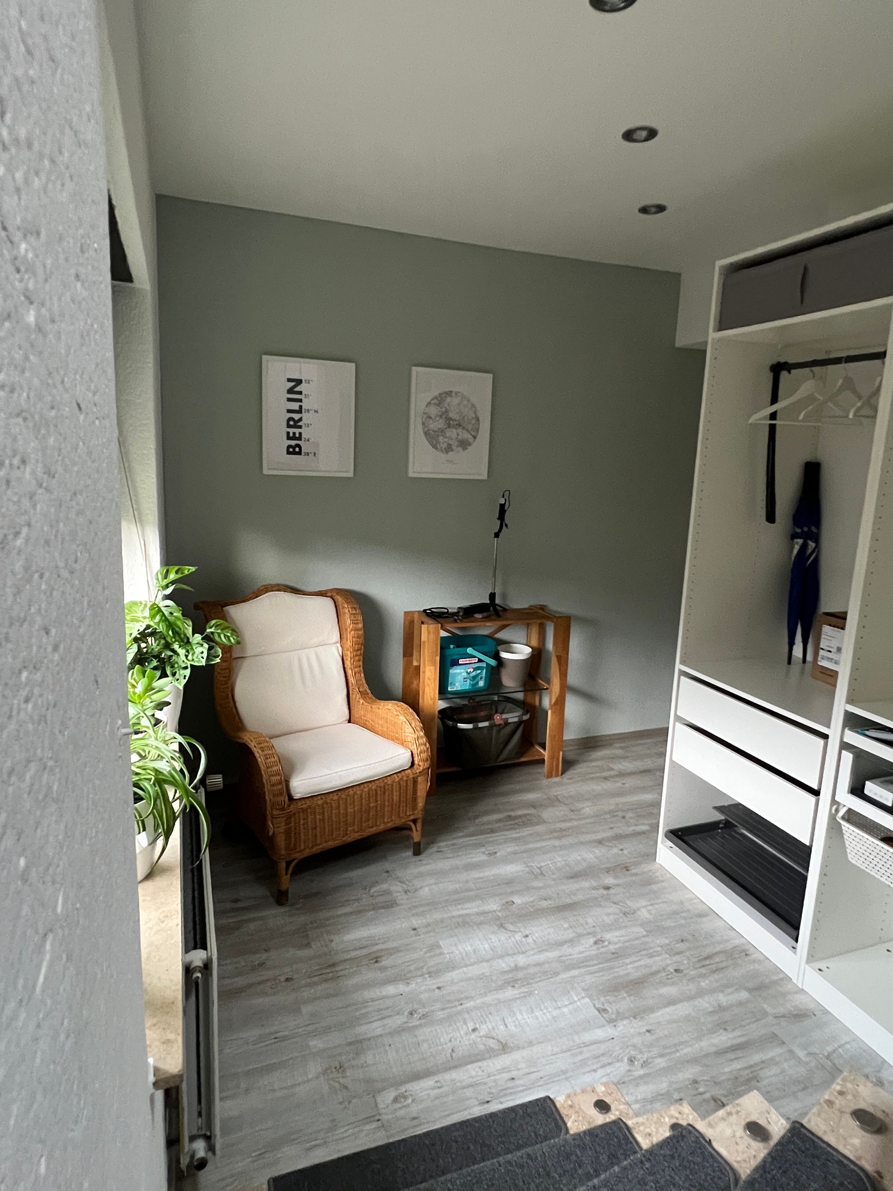 A room with a wicker chair, a small wooden table with a lamp, a white wardrobe, and two framed pictures on the wall.