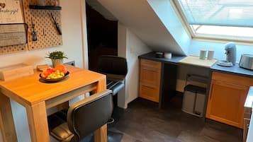 Eine kleine Küche mit Holztisch, schwarzen Stühlen, einer Arbeitsplatte und einem Fenster mit Jalousien.