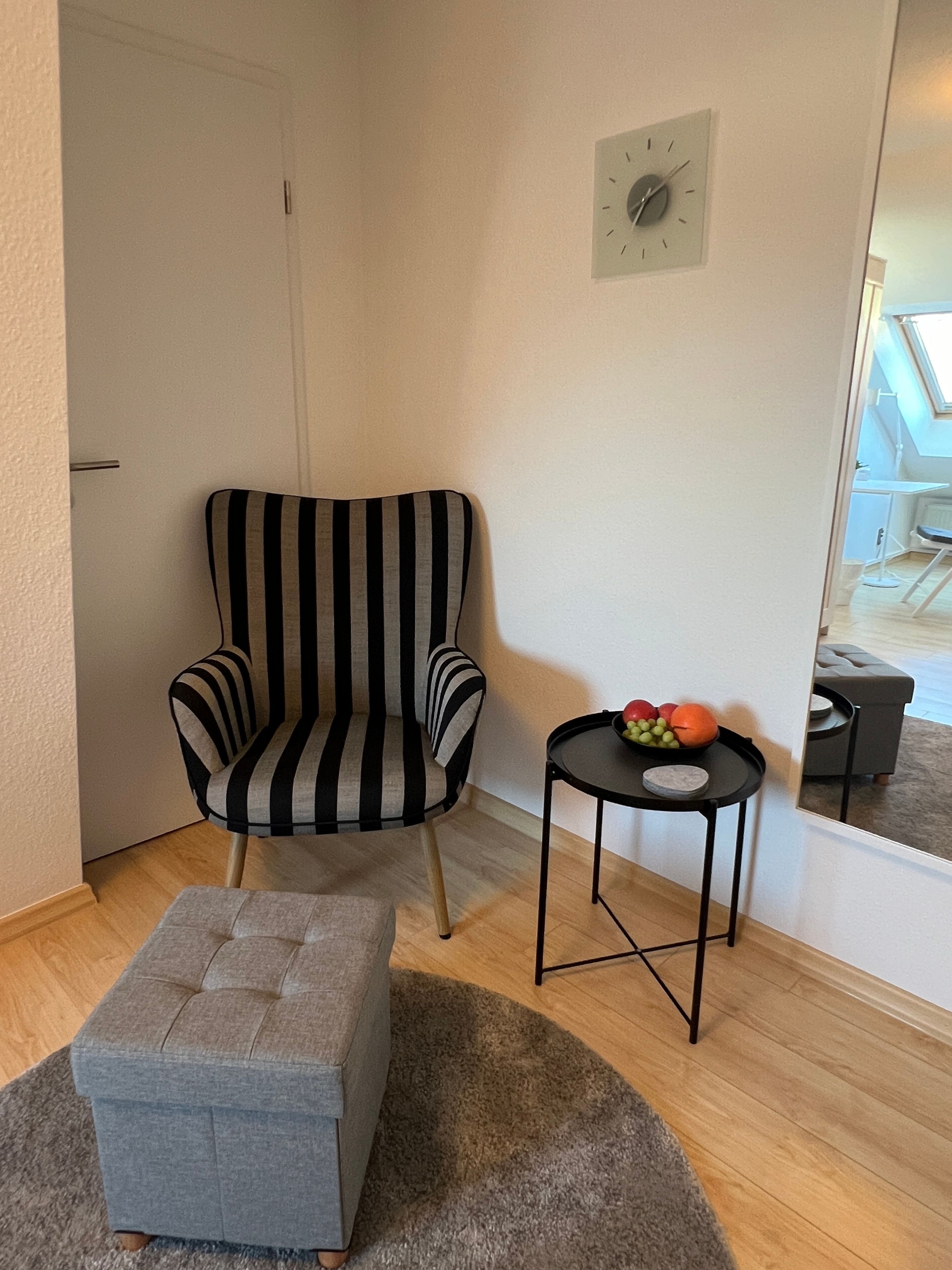 Ein Eckzimmer mit einem gestreiften Sessel, einem kleinen runden Tisch mit Obst, einem grauen Hocker und einer Wanduhr.