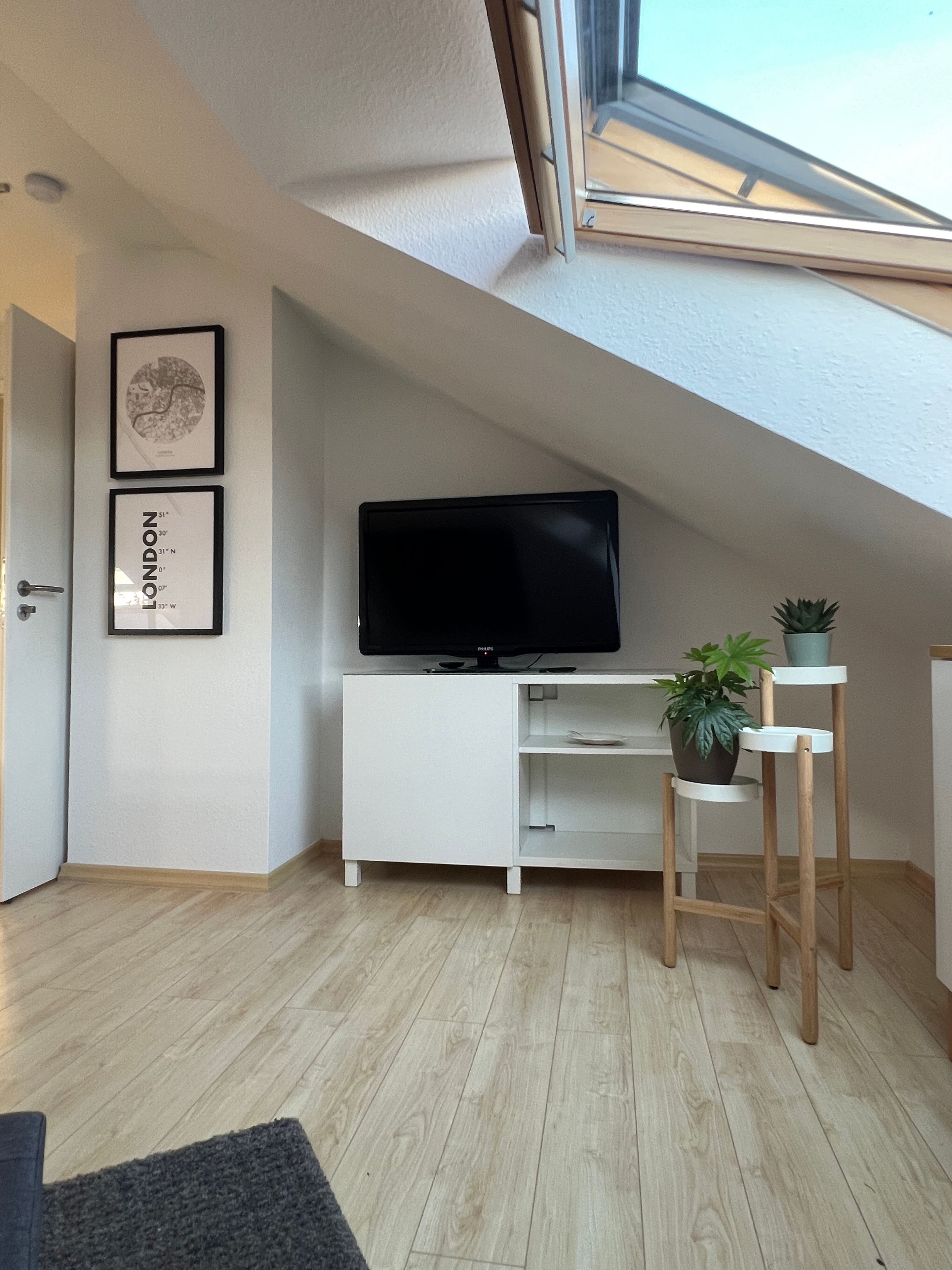 Ein modernes Wohnzimmer mit schräg versiegelter Decke, einem Flachbildfernseher auf einer weißen Kommode, einem kleinen Holzschemel mit Pflanze und zwei gerahmten Bildern an der Wand.