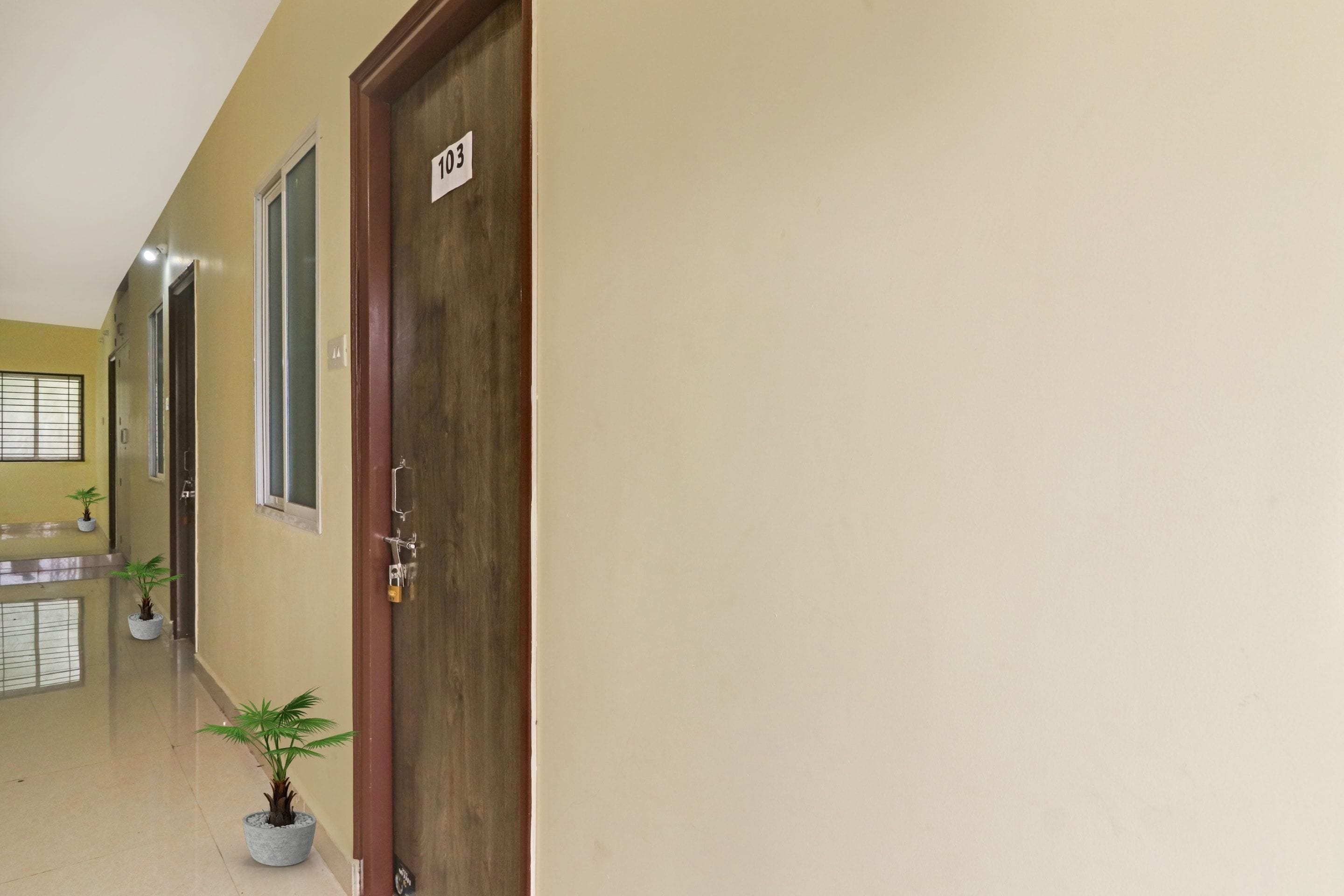 Un pasillo con puerta de madera marcada '103', plantas en macetas y piso de baldosas.