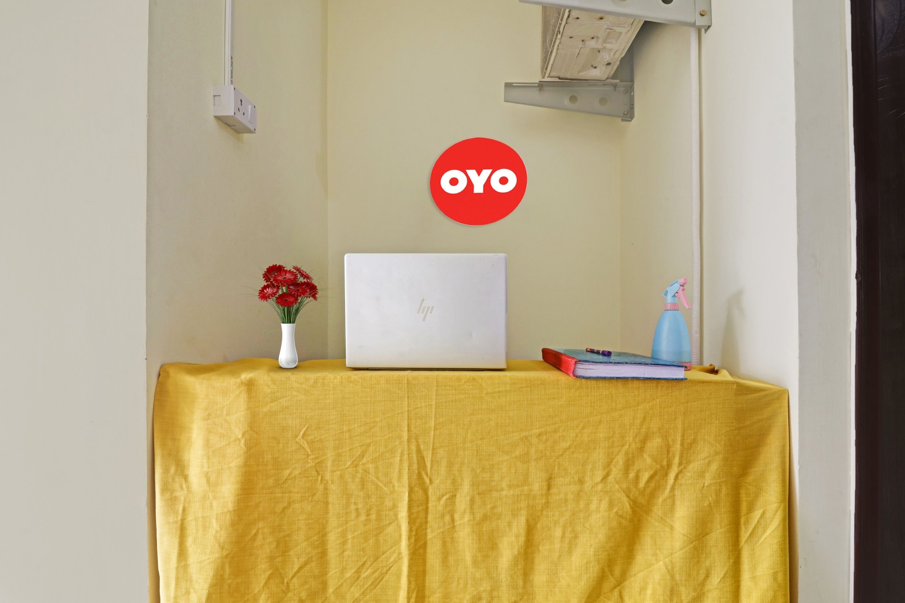 A laptop on a yellow table with a red flower vase, a book, and a water bottle.