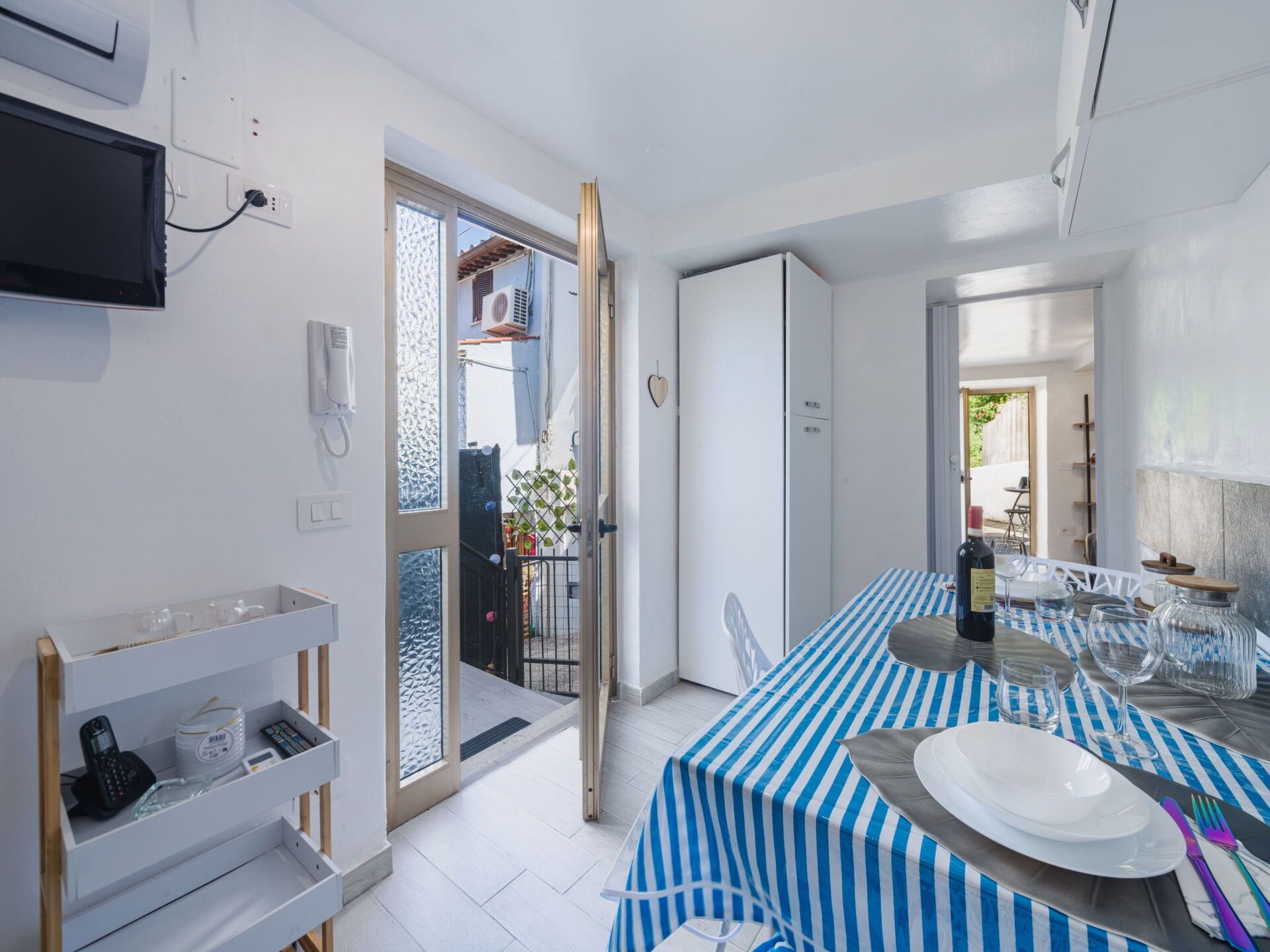 A modern kitchen with a dining area, a TV mounted on the wall, and a door leading to a balcony.