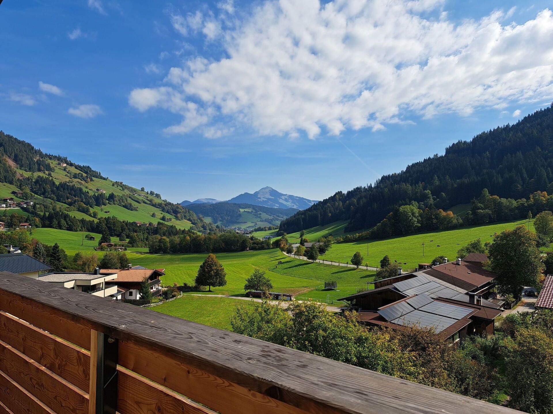 A view from a balcony overlooking a valley with houses, green fields, and mountains in the distance.