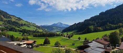 A view from a balcony overlooking a valley with houses, green fields, and mountains in the distance.