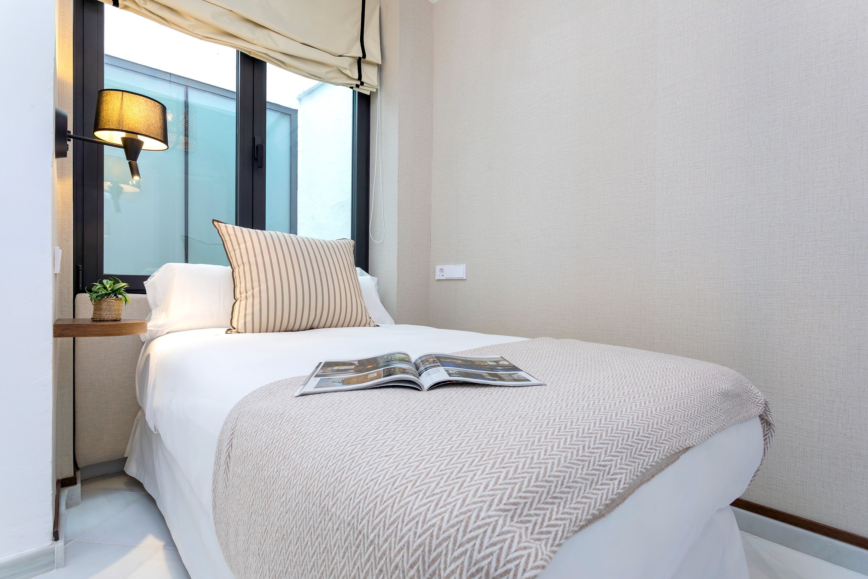 A neatly made bed with a magazine, a lamp, and a plant on a bedside table.