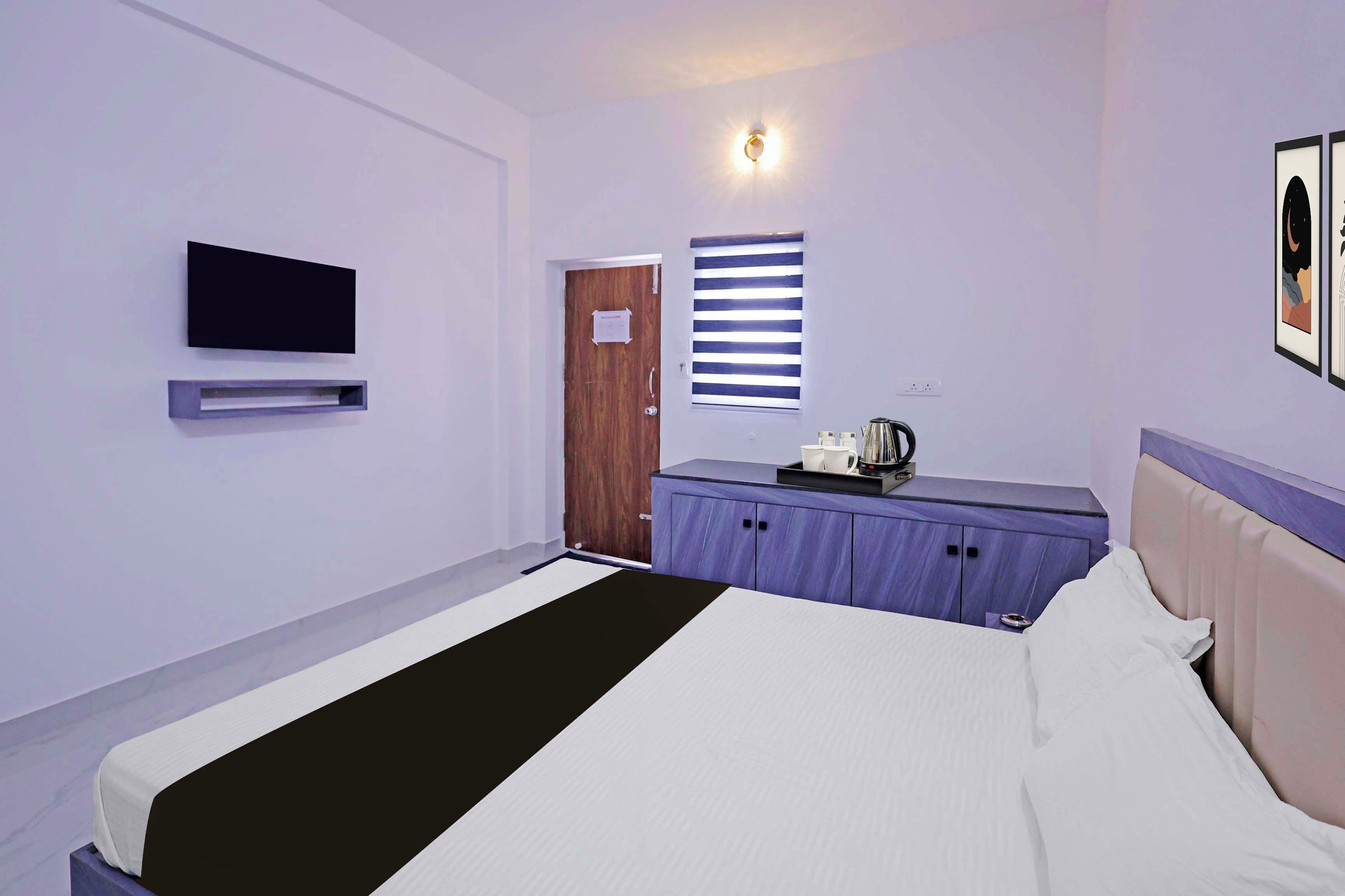 Une chambre d’hôtel avec un lit, un téléviseur fixé au mur, une fenêtre avec des stores, une porte et une petite table sur laquelle se trouvent une bouilloire et des tasses.