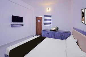 A hotel room with a bed, a television mounted on the wall, a window with blinds, a door, and a small table with a kettle and cups.