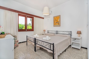 Ein Schlafzimmer mit einem Bett, zwei Nachttischen, einem Fenster mit Vorhängen, einem Bild an der Wand und einem kleinen Tisch mit einer Lampe.