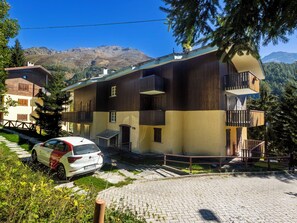 Ein zweigeschossiges Gebäude mit einem Balkon im oberen Stockwerk, ein Auto vor der Tür und Berge im Hintergrund.