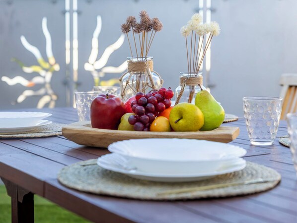 Une table de pique-nique en bois, agrémentée d’un centre de table composé de fruits et d’un vase en verre, entourée d’assiettes et de verres.
