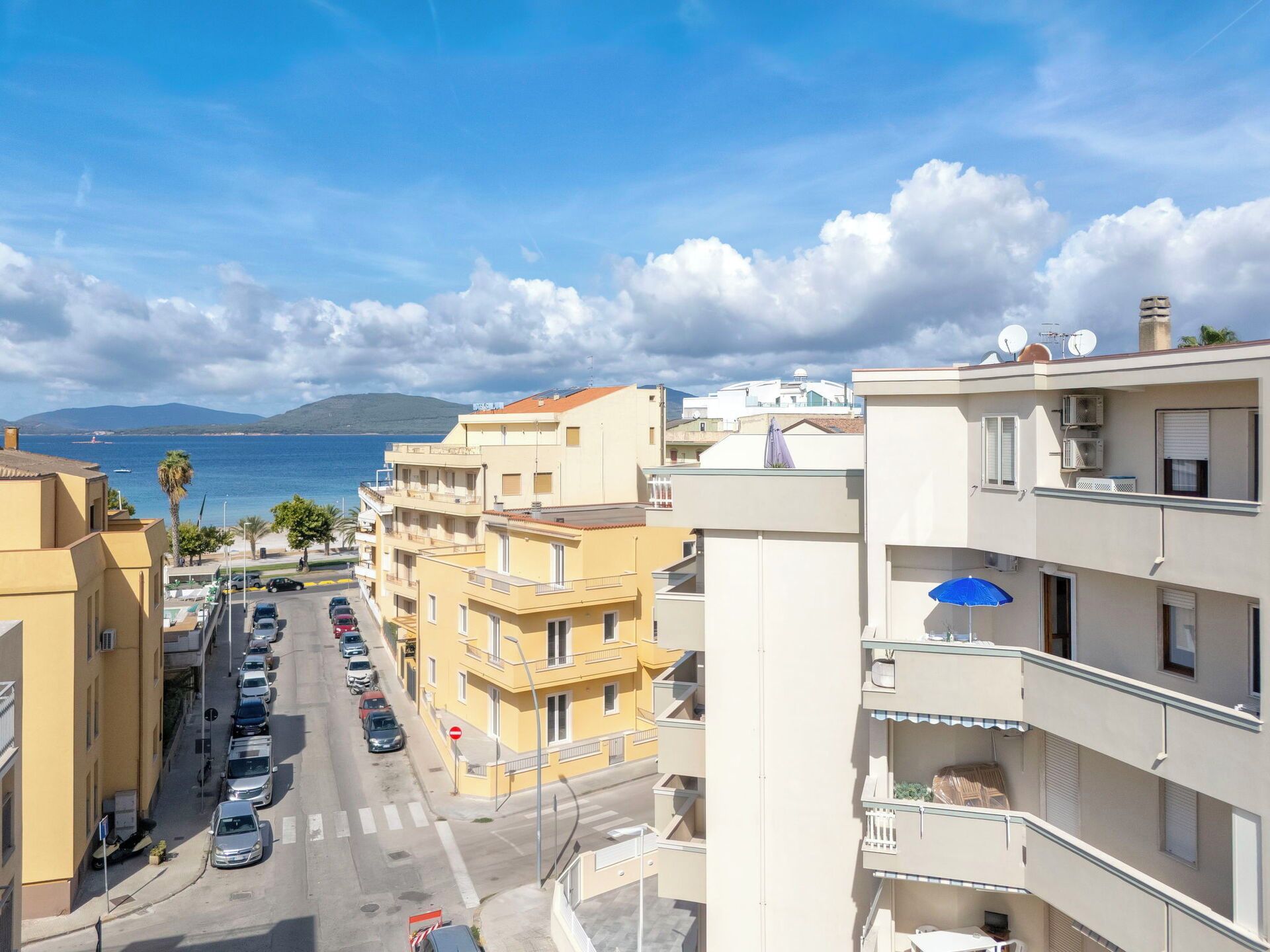 A coastal residential area with multi-story buildings, a street with parked cars, and a view of the sea and mountains in the distance.