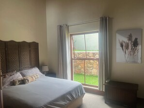 A bedroom with a large bed, a headboard, a nightstand, a lamp, a window with curtains, a view of the outdoors, and a wall-mounted artwork. - Private Pretoria Villa for 12 with Pool, Cinema, Bar & Firepit (Pretoria)