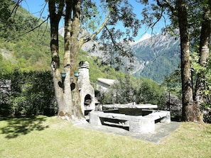 A stone structure with a bench and a small building, surrounded by trees and mountains. - Ca' Angiulin by Interhome (Olivone)