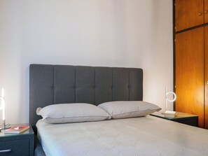 A neatly made bed with a grey upholstered headboard, a bedside table with a lamp, and a wooden wardrobe. - Ca' Angiulin by Interhome (Olivone)