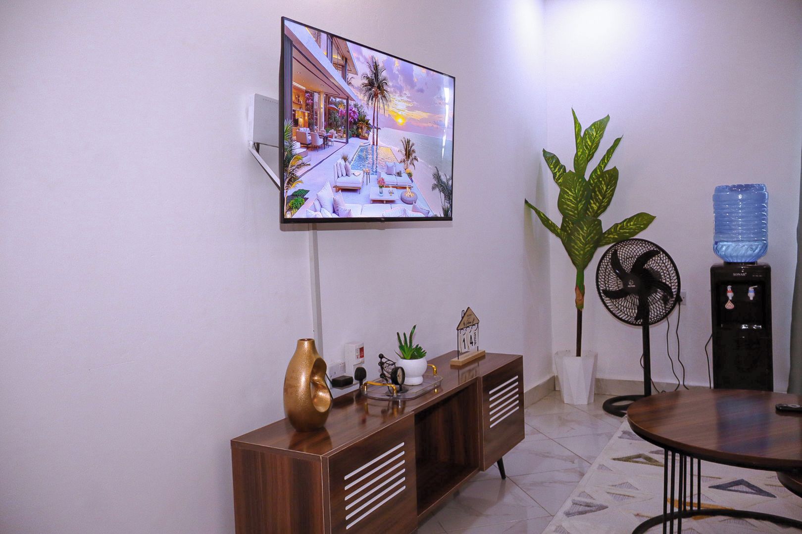 A modern living room with a flat-screen TV mounted on the wall, a wooden console table with decorative items, a water dispenser, a large potted plant, and a standing fan.